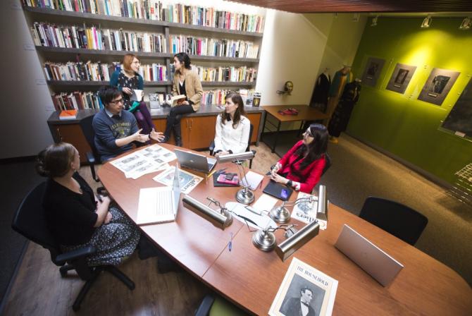 Au premier plan, 4 personnes sont assises et discutent à une table de réunion couverte de papiers, de stylos et d’ordinateurs portables; en arrière-plan, 2 personnes sont assises et discutent devant une grande bibliothèque, chacune tenant un livre ouvert