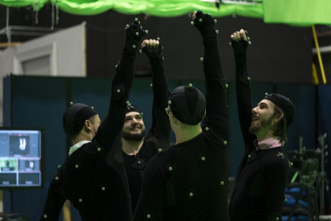 Four people dressed with black body suits and caps covered with small green balls stand in a circle, each of them raising an arm