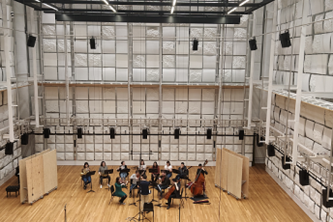 An orchestra performs in a large room where walls are covered with panelling and speakers
