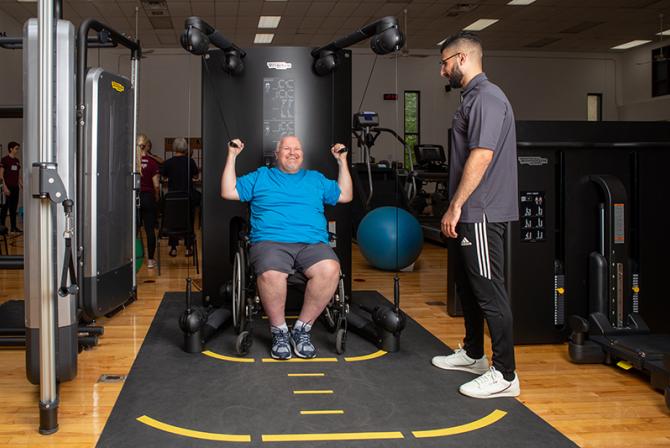 A person in a wheelchair uses equipment to exercise their arms as another supervises them.