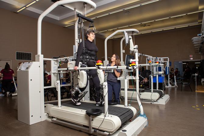 A person wears a robotic assist walking device while using a treadmill as another person supervises them.