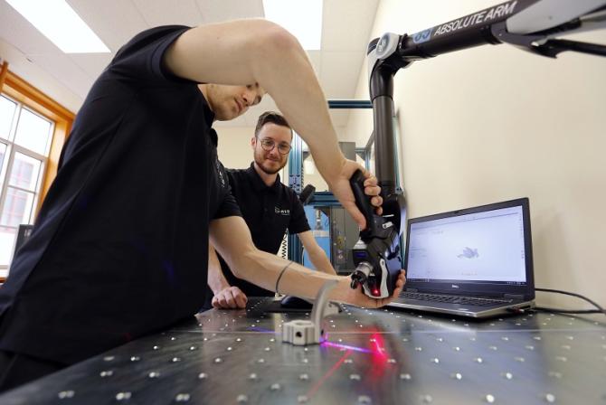 A person manipulates a robot arm over a part as another person looks on.