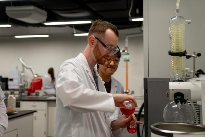 A researcher pours red liquid from a flask into a smaller one.
