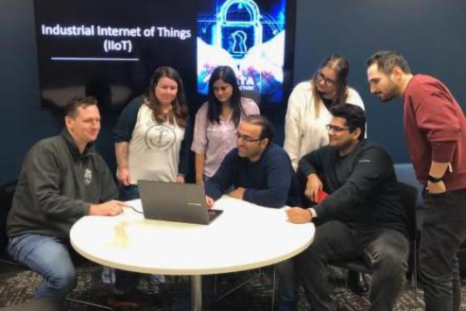Un groupe de personnes entoure une personne assise qui travaille sur un ordinateur portable. Un écran derrière eux affiche une diapositive intitulée "Industrial Internet of Things (IoiT)".