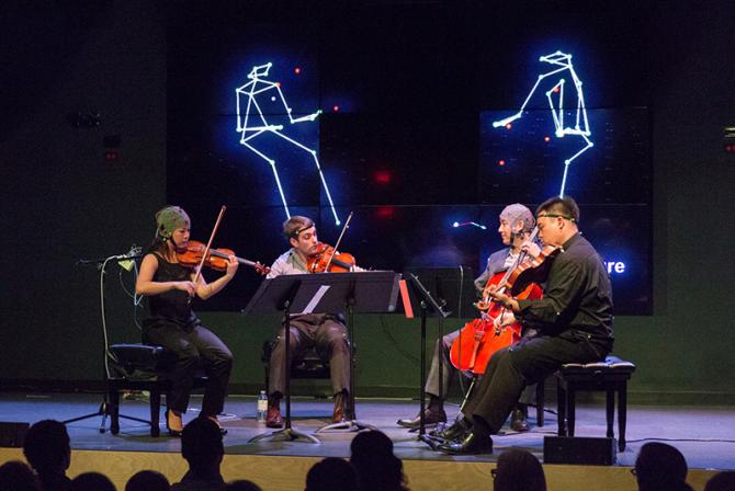 Musicians wear tracking devices as they perform on stage. A graphic representation of their movements displays on a large screen behind them.