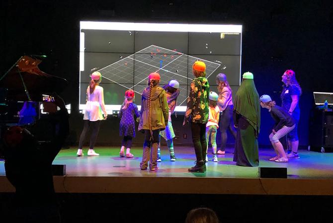 A group of people wearing caps with sensors face a large screen on a stage.