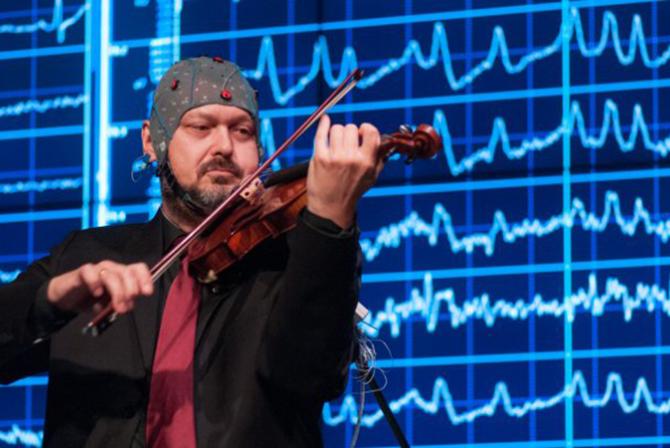 A musician wearing an EEG cap plays the violin in front of a large screen displaying wavy lines.